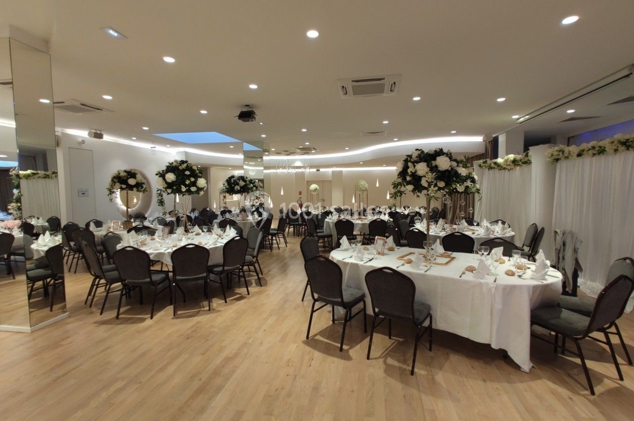 Salle de réception élégante avec tables rondes dressées, décorations florales blanches et éclairage chaleureux.