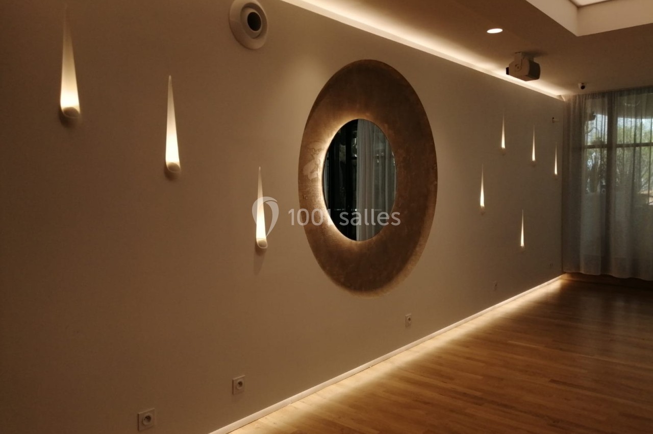Mur beige avec un miroir rond central, éclairages muraux en forme de gouttes et sol en parquet clair.