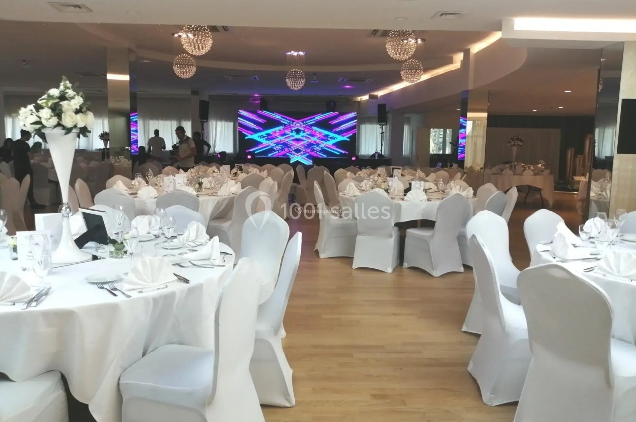Salle de réception élégante avec tables rondes dressées, chaises blanches et écran lumineux en arrière-plan.