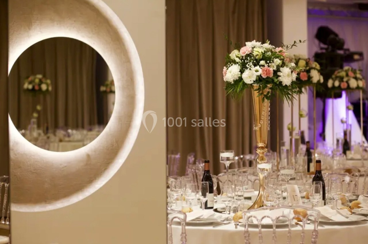 Salle de réception élégante avec tables dressées, décor floral sur pied doré et éclairage tamisé.