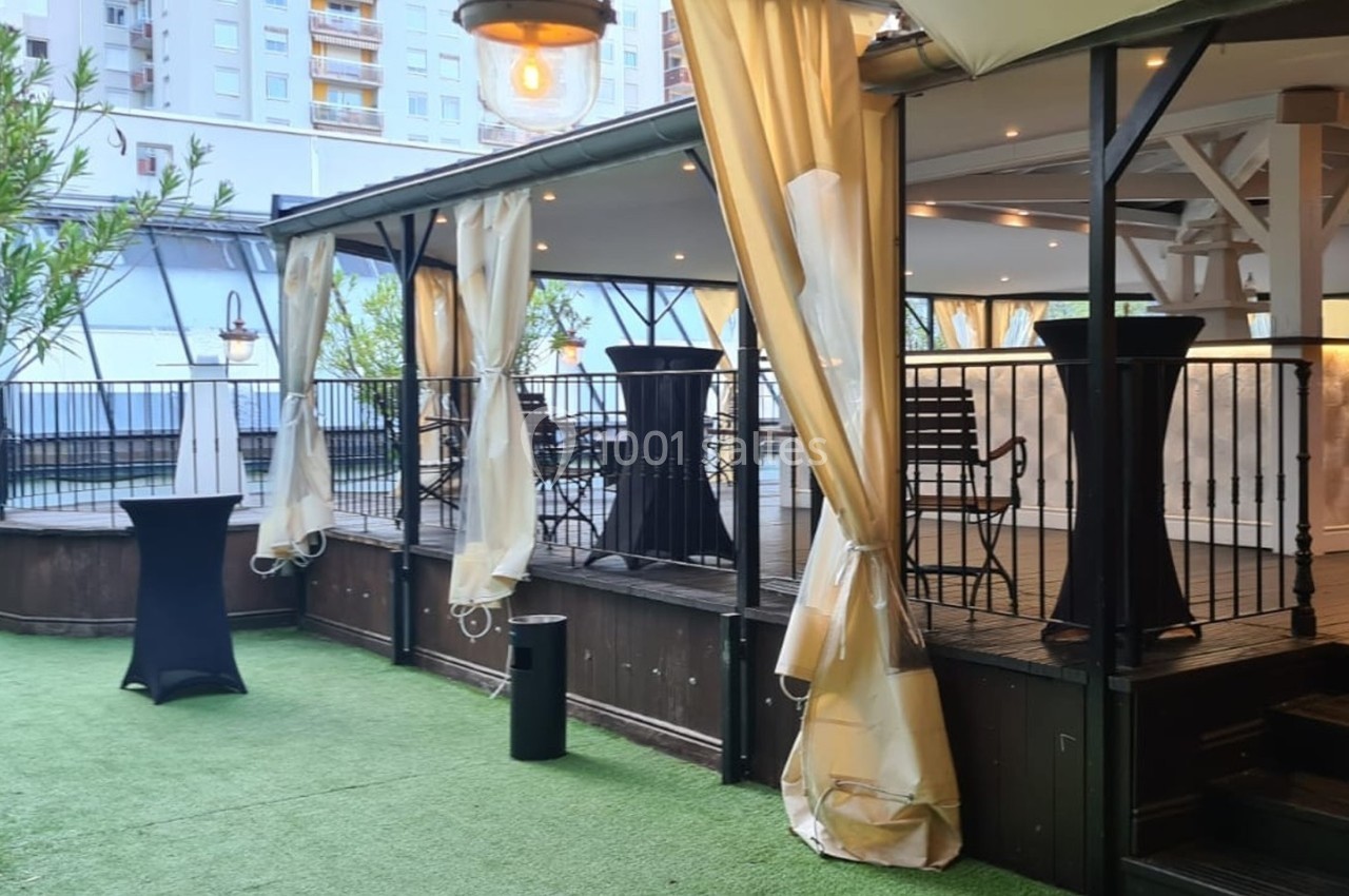 Terrasse aménagée avec tables hautes et rideaux, entourée de verdure et située près d'immeubles modernes.