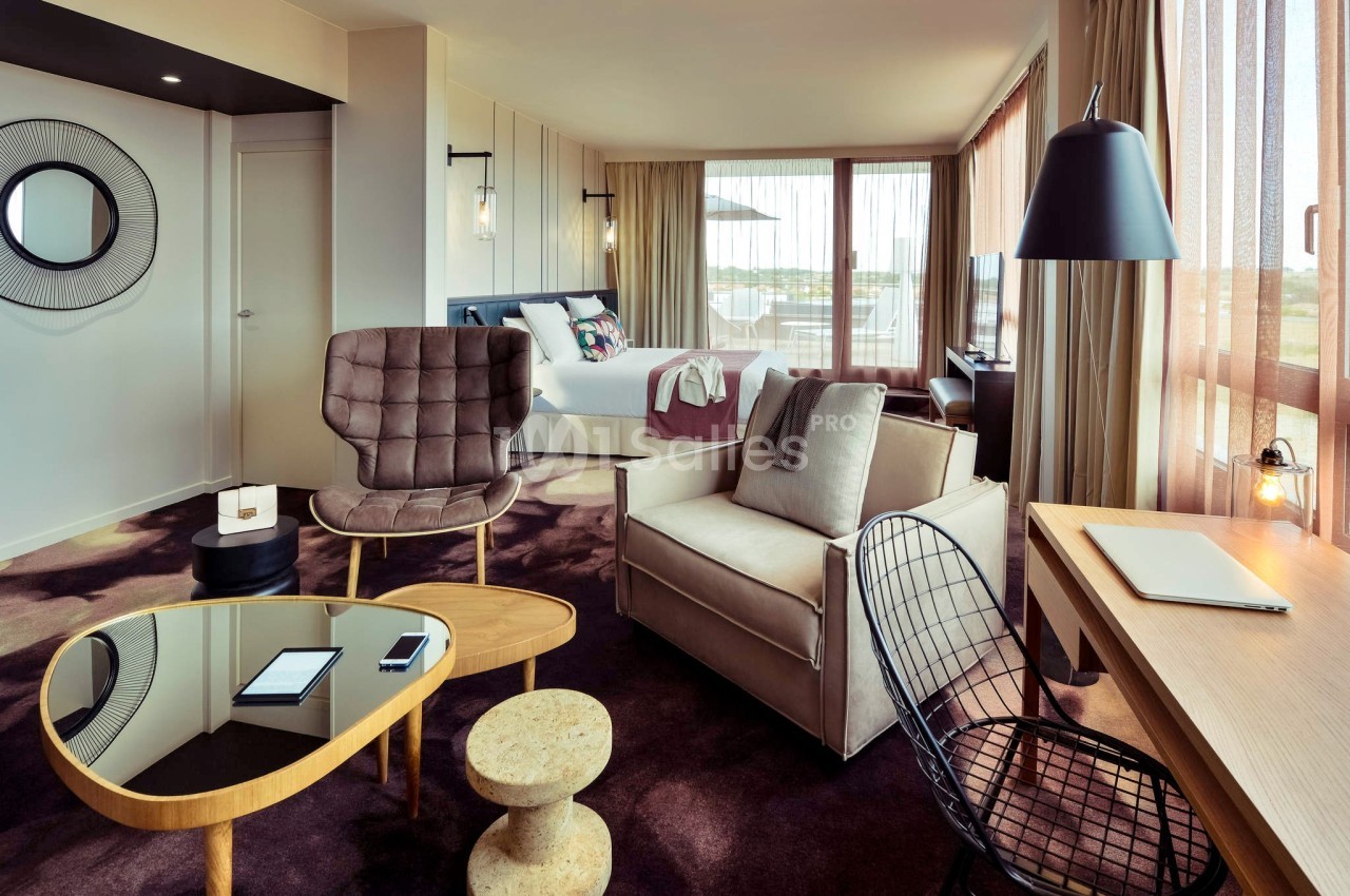 Chambre d'hôtel moderne avec lit, coin salon, bureau en bois et grandes fenêtres laissant entrer la lumière naturelle.