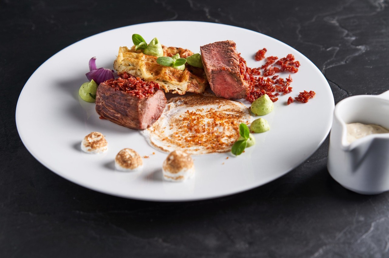 Assiette gastronomique avec viande grillée, purée verte, chips, sauce blanche et décorations raffinées sur une table noire.