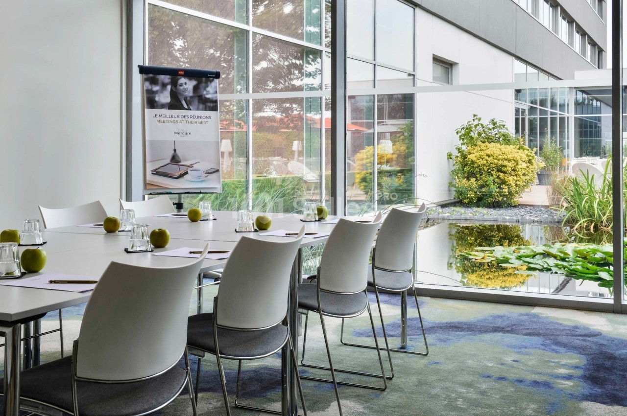 Salle de réunion lumineuse avec table rectangulaire, chaises blanches, pommes vertes et vue sur un jardin extérieur.
