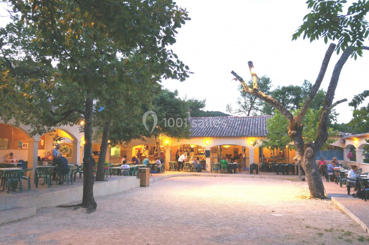 Terrasse extérieure d'un restaurant éclairée en soirée, entourée d'arbres et occupée par des clients.