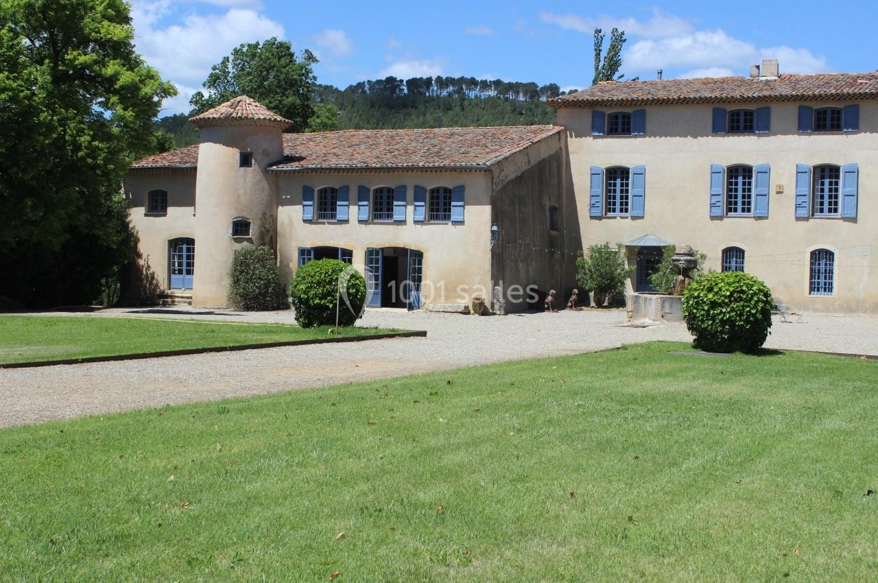 Bâtiment ancien en pierre avec volets bleus, entouré de pelouses et d'arbres sous un ciel ensoleillé.