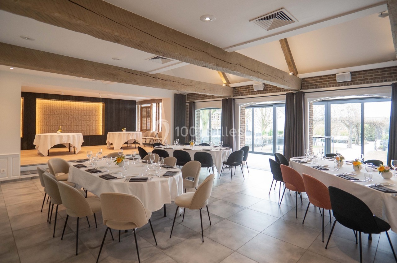 Salle lumineuse aménagée pour un événement, avec tables rondes dressées, chaises variées et grandes fenêtres donnant sur l…