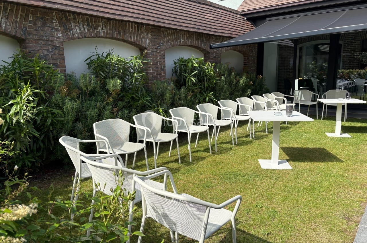 Chaises blanches disposées autour de tables sur une pelouse, près d'un bâtiment en briques et entourées de végétation.