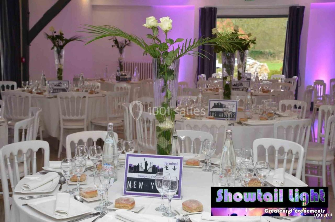 Salle de réception décorée avec des tables rondes, nappes blanches, centres de table floraux et éclairage violet.