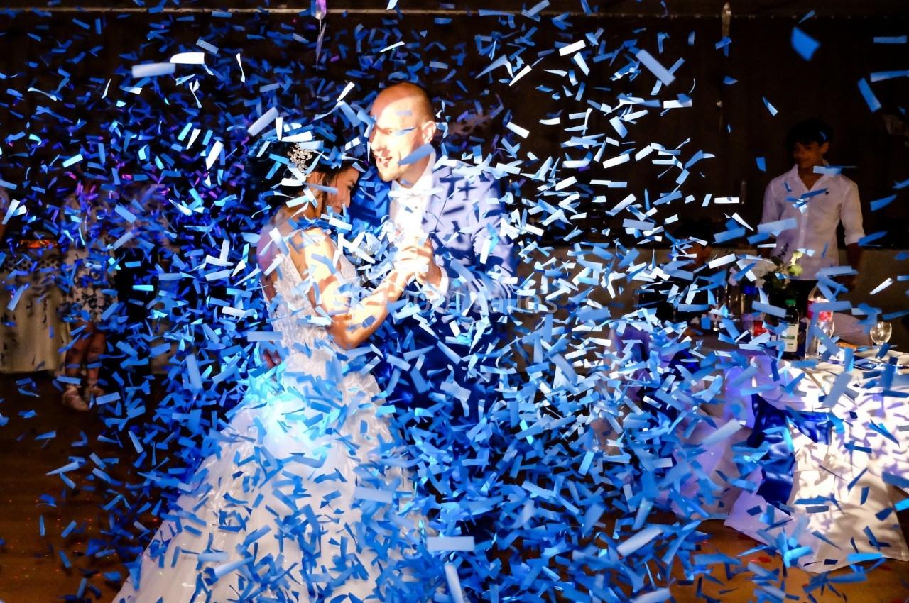 Un couple danse au milieu de confettis bleus dans une salle de réception faiblement éclairée.