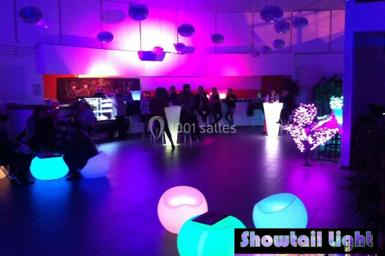 Salle éclairée par des lumières colorées avec des tables hautes, des sièges lumineux et un groupe de personnes en arrière…