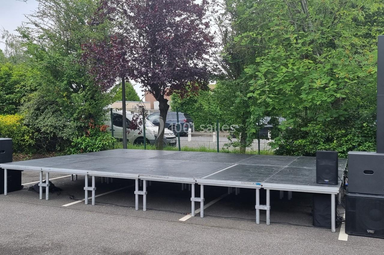 Scène extérieure montée sur un parking, entourée de végétation et équipée de haut-parleurs.