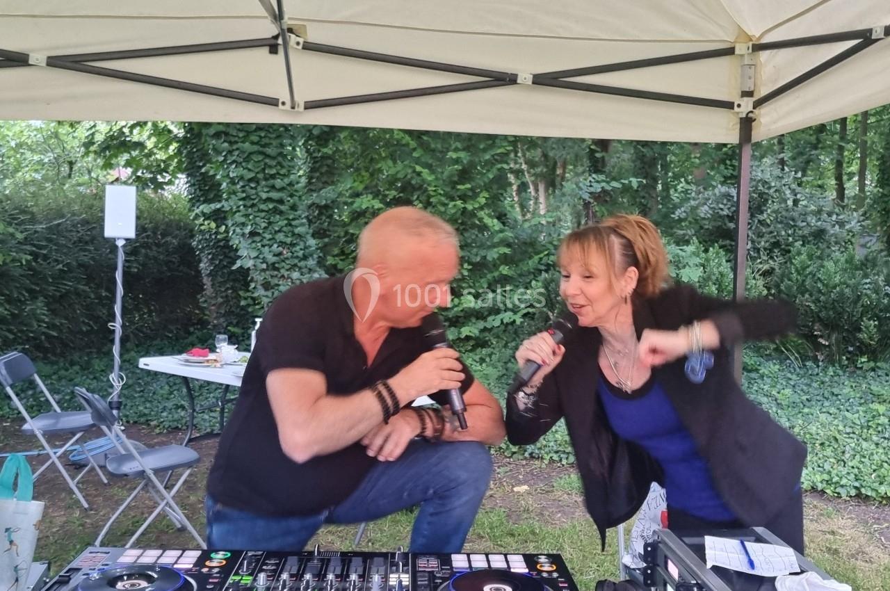 Un homme et une femme chantent ensemble sous une tente, près d'une table de mixage, dans un cadre boisé.