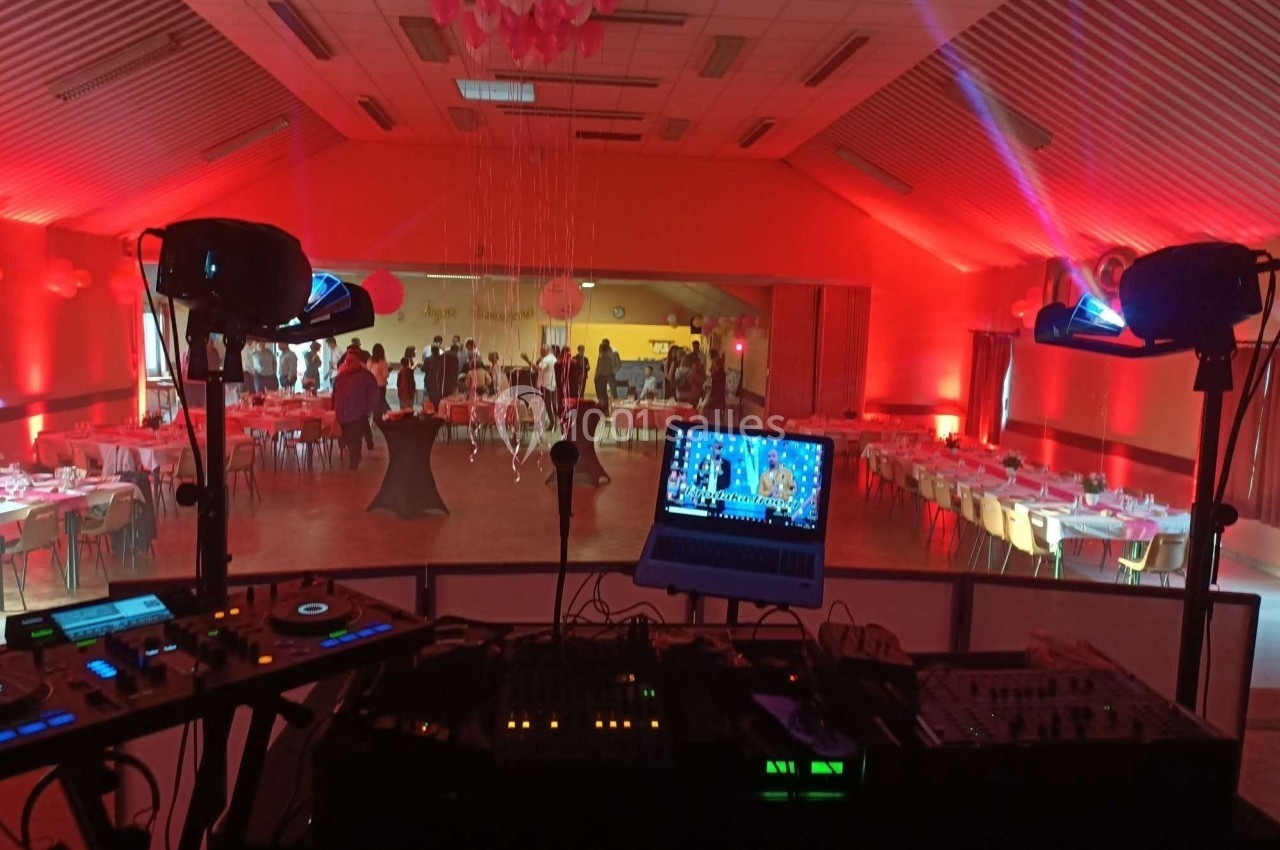 Salle de réception éclairée en rouge, vue depuis une table de mixage avec des invités et des tables dressées au fond.