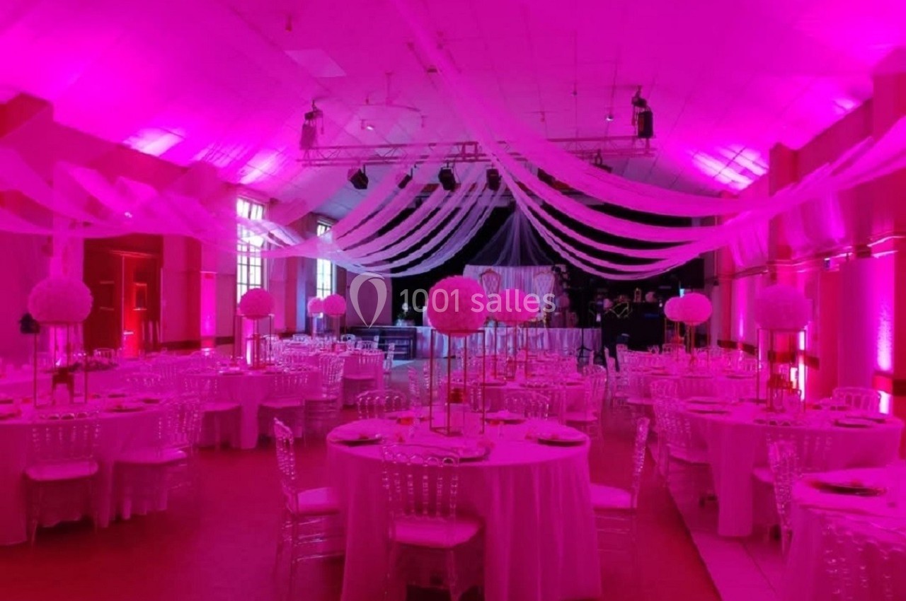 Salle de réception décorée avec des tables rondes, nappes blanches, chaises transparentes et éclairage rose.
