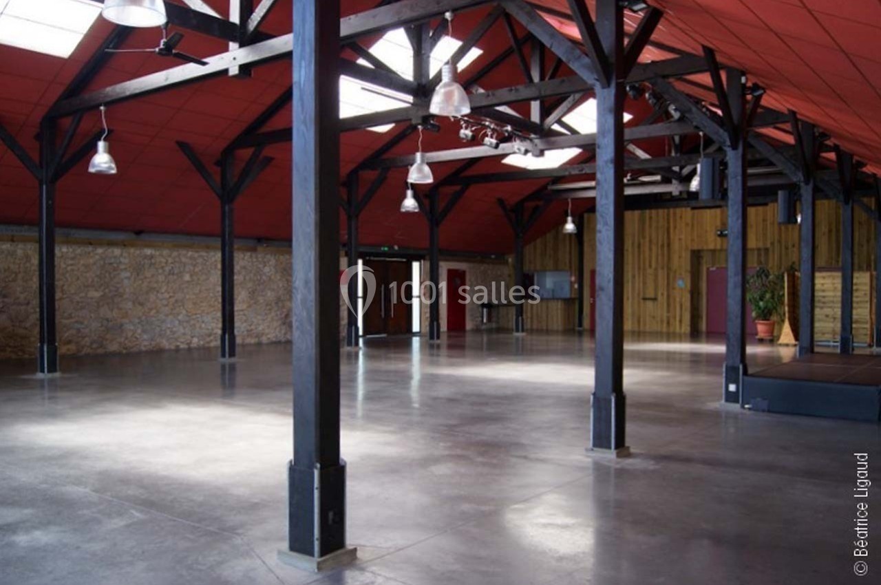 Salle spacieuse avec poutres apparentes, murs en pierre et bois, sol en béton lisse et éclairage suspendu.
