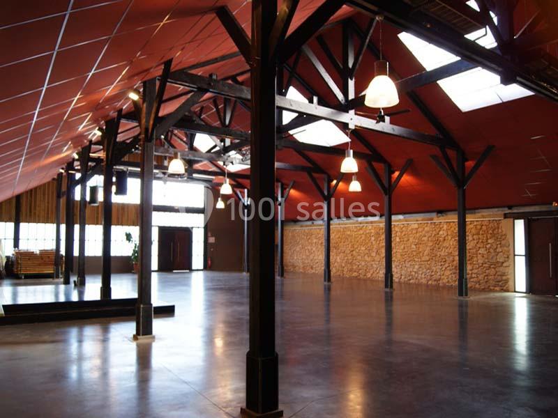 Intérieur d'une grande salle avec charpente en bois, murs en pierre, sol en béton et plafond rouge avec luminaires suspendus.
