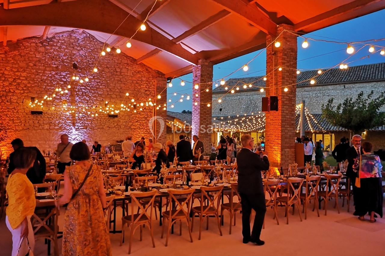 Salle de réception en soirée avec tables dressées, invités debout et guirlandes lumineuses suspendues.