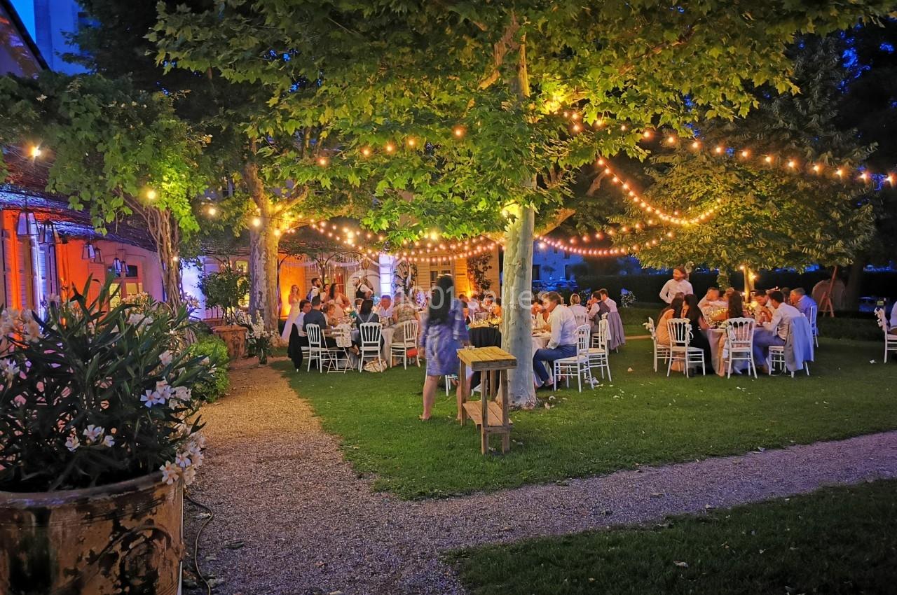 Dîner en plein air sous des guirlandes lumineuses, avec des invités assis à des tables dans un jardin en soirée.