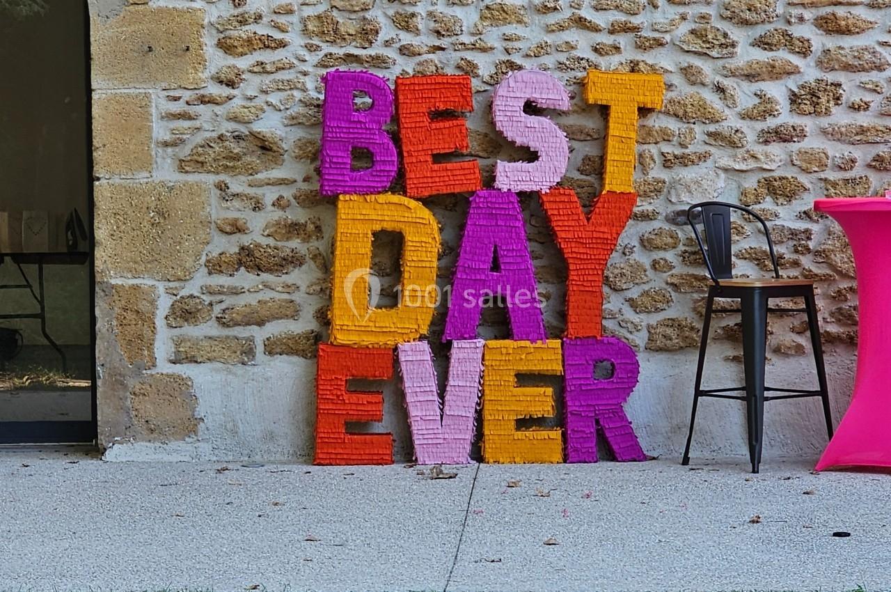 Lettres colorées formant ’Best Day Ever’ appuyées contre un mur en pierre, près d'une table et d'une chaise.