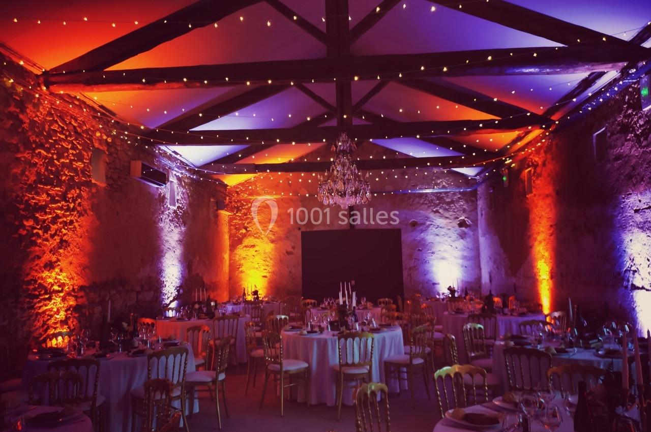 Salle de réception en pierre éclairée par des lumières colorées, avec tables rondes dressées pour un événement.