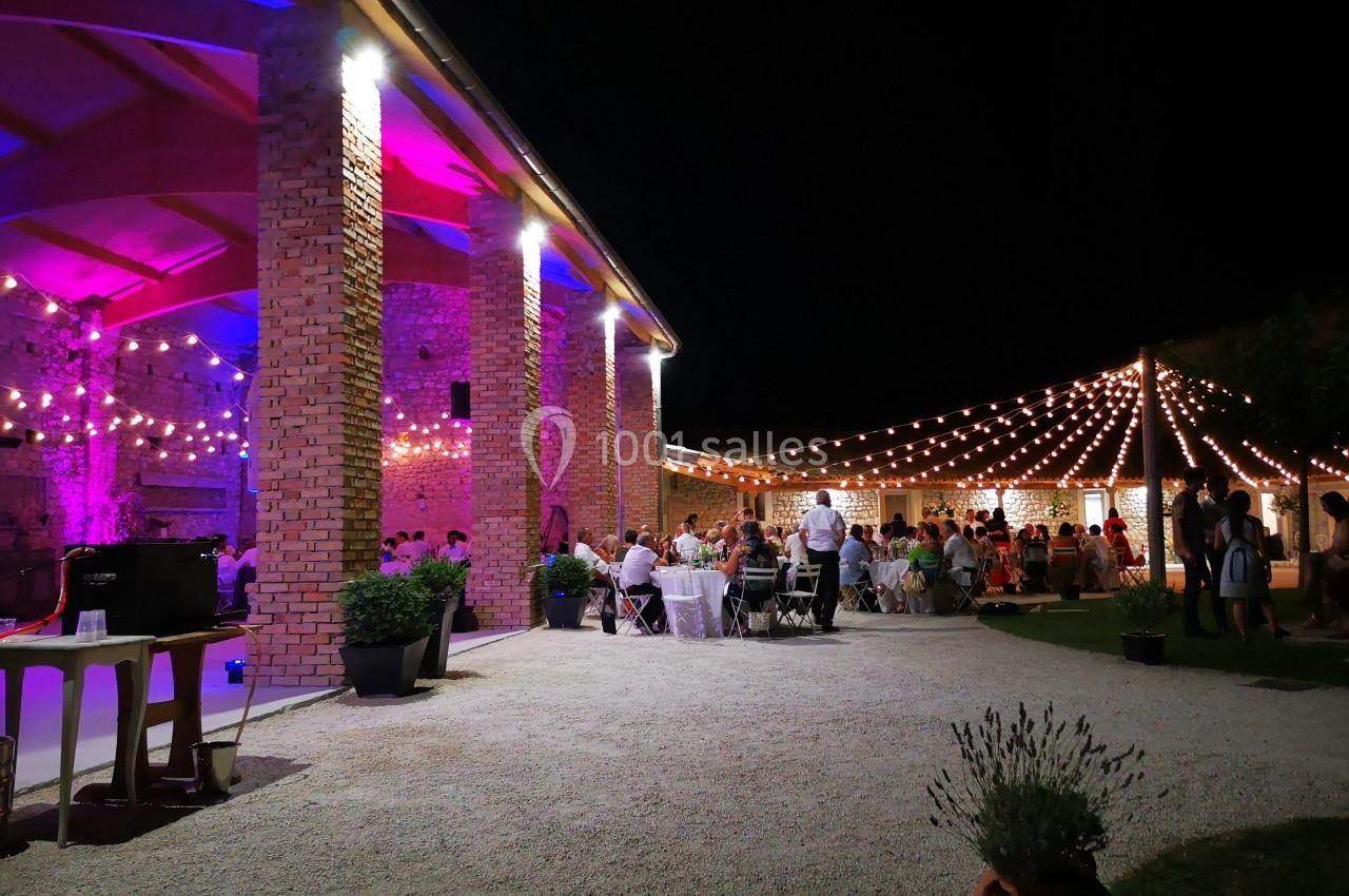 Cour extérieure éclairée par des guirlandes lumineuses, avec des invités assis à des tables lors d'un événement nocturne.