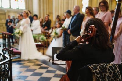 Miniature Violoncelle Une violoncelliste joue lors d'une cérémonie dans une église, entourée d'invités debout et de mariés à l'arrière-plan.