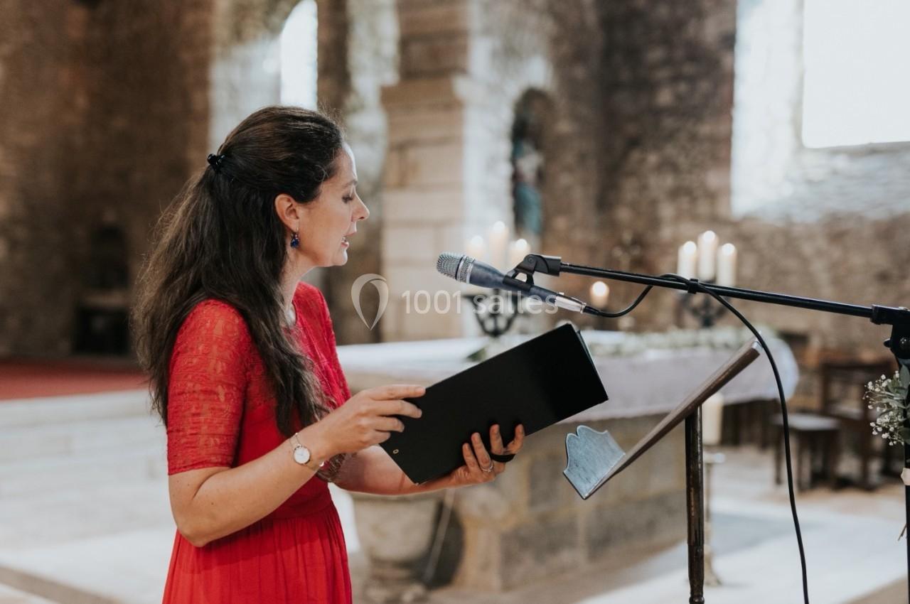 Femme en robe rouge lisant un texte devant un micro dans une église en pierre éclairée par la lumière naturelle.