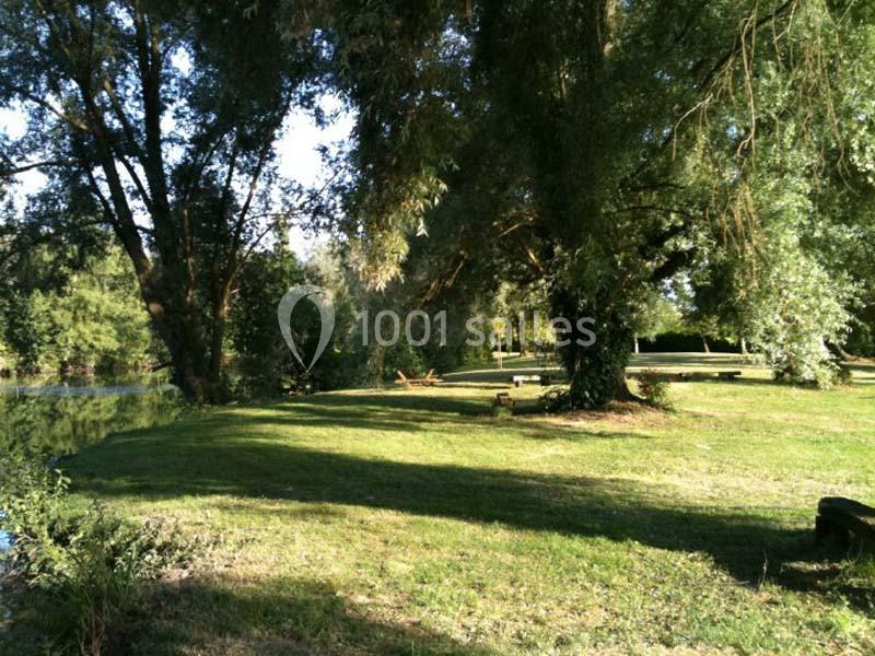 Un parc verdoyant avec de grands arbres, une pelouse ombragée et un cours d'eau visible à gauche.