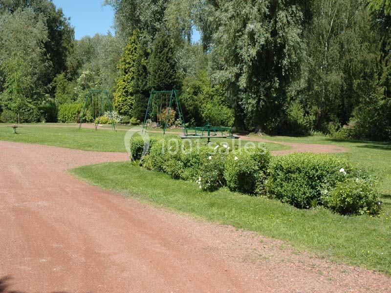 Allée en gravier bordée de pelouses et arbustes, avec des balançoires et des arbres en arrière-plan dans un parc.