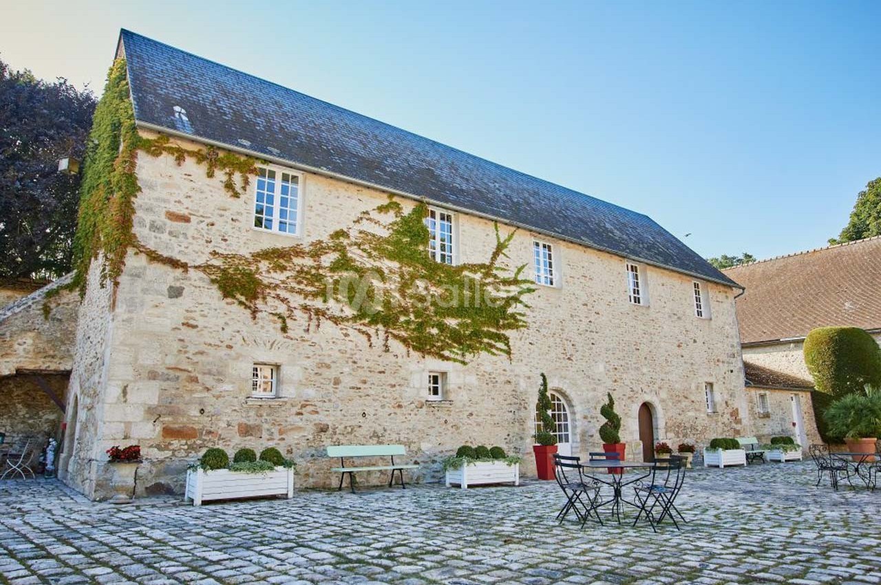 Bâtisse en pierre avec toit en ardoise, façade partiellement couverte de lierre, entourée d'une cour pavée.