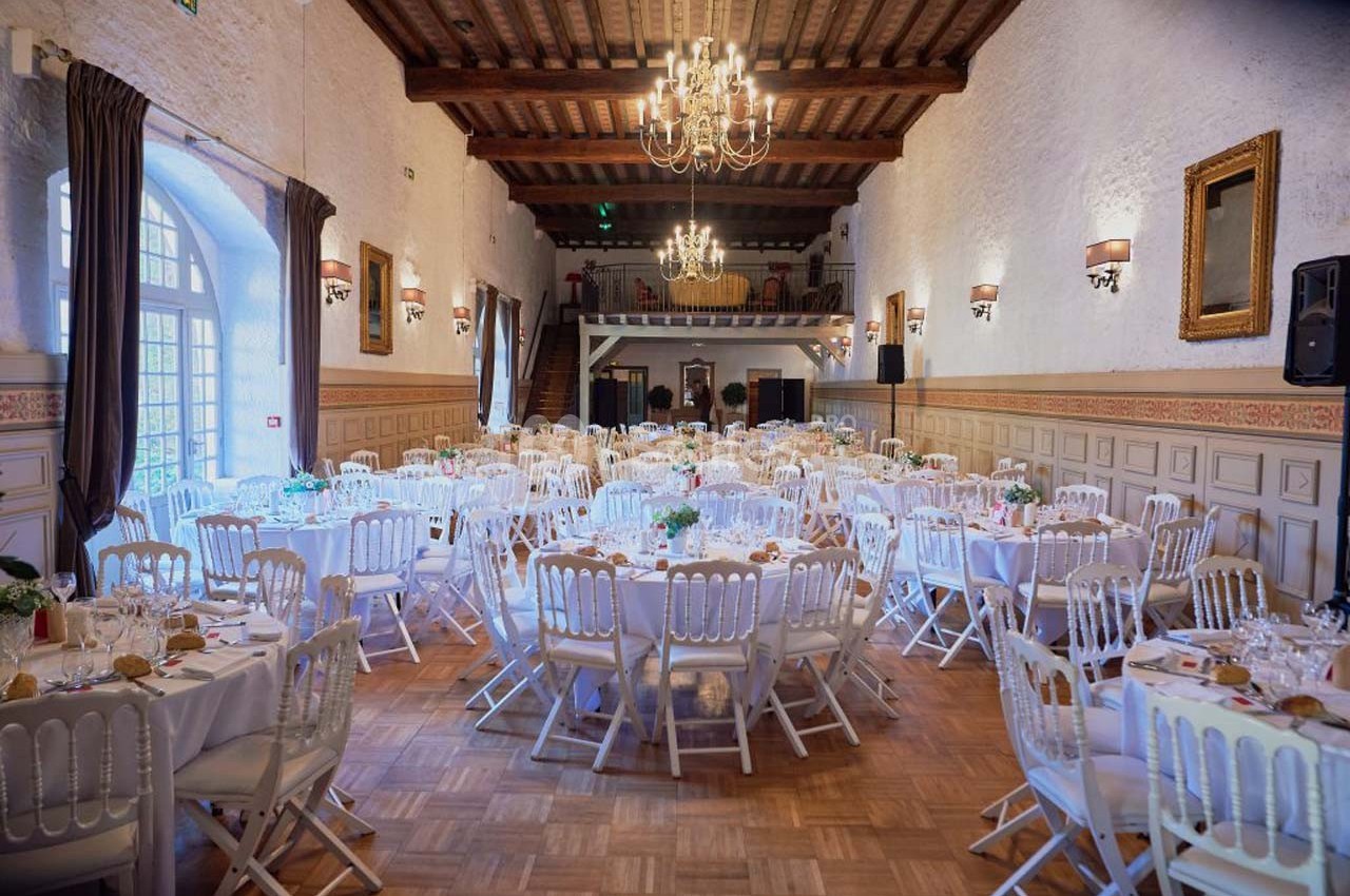 Salle de réception élégante avec tables rondes dressées, chaises blanches et lustres suspendus au plafond en bois.