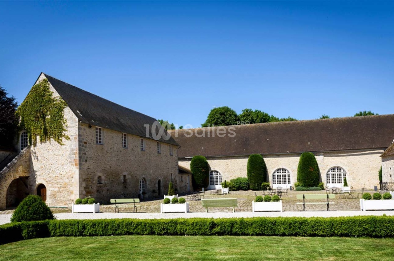 Cour intérieure d'un bâtiment en pierre avec des fenêtres cintrées, des bancs et des haies taillées sous un ciel bleu.