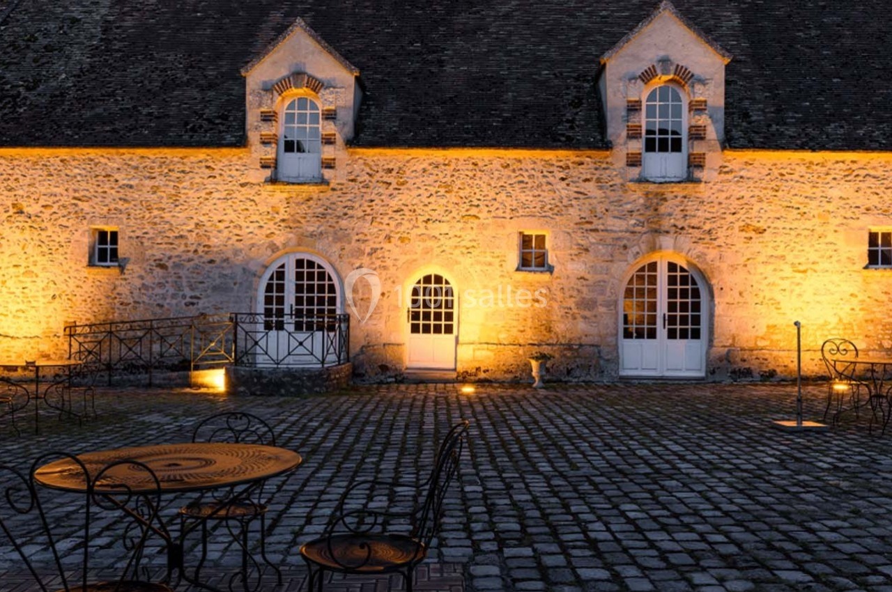 Cour pavée avec des tables en fer forgé, devant une bâtisse en pierre éclairée par des lumières chaudes.
