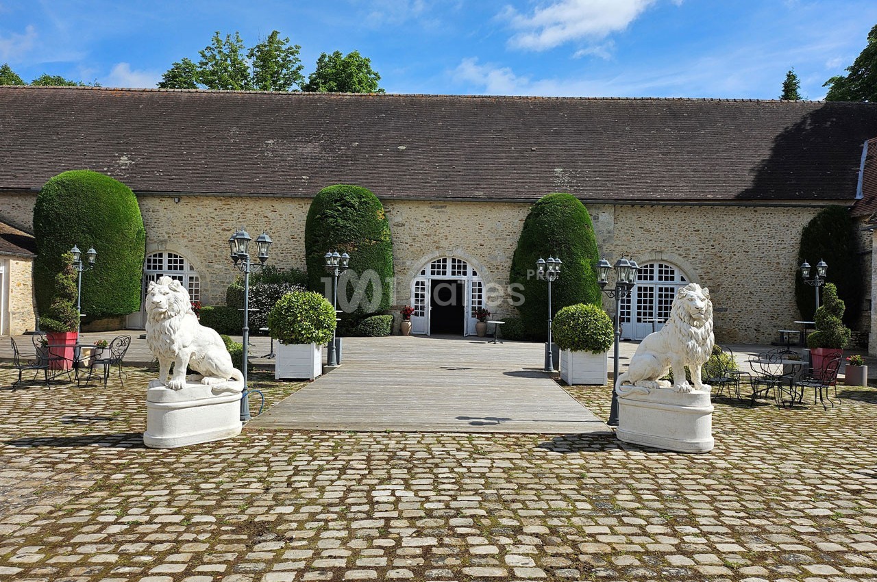 Cour pavée avec deux statues de lions en pierre encadrant une entrée voûtée d'un bâtiment en pierre avec toit en ardoise.
