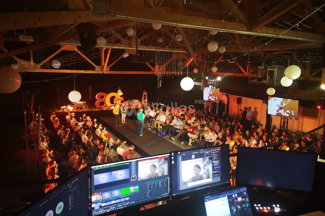 Salle comble avec public assis, scène éclairée, écrans de contrôle au premier plan et décorations suspendues.