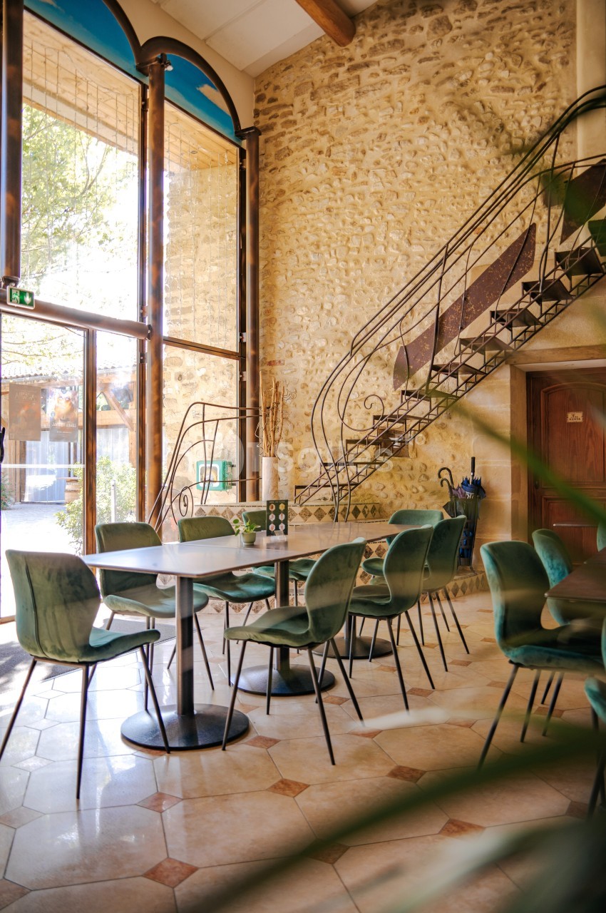 Salle lumineuse avec murs en pierre, escalier en fer forgé, grandes baies vitrées et chaises vertes autour de tables.