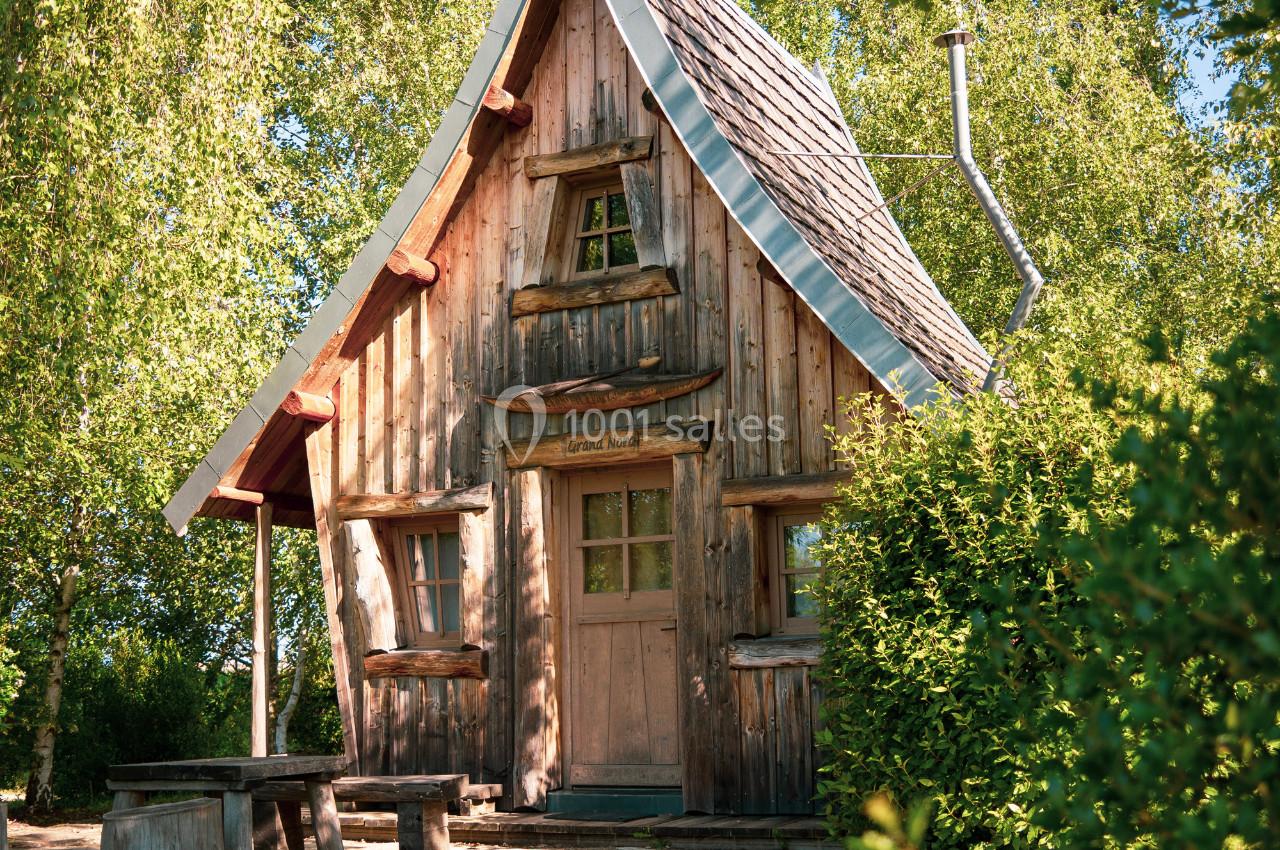 Cabane en bois avec toit pentu entourée de verdure, vue ensoleillée avec une table en bois à l'avant.