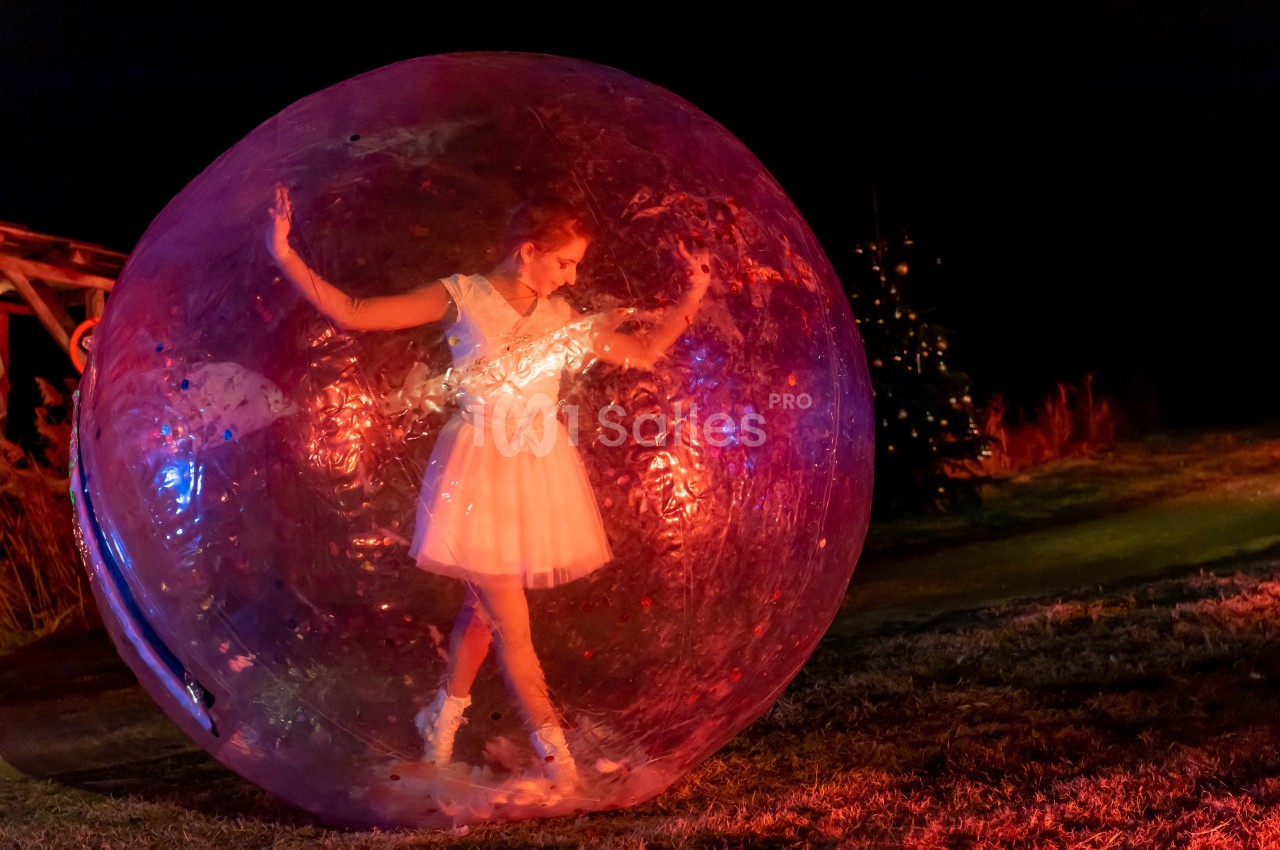 Femme en robe blanche dans une grande bulle transparente, éclairée par des lumières colorées dans un environnement nocturne.
