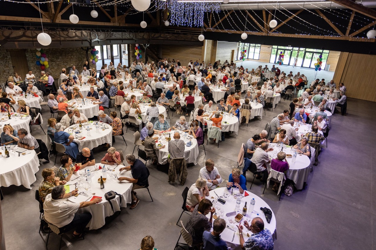 Salle de réception remplie de convives assis à des tables rondes, décorée de guirlandes lumineuses et de ballons.