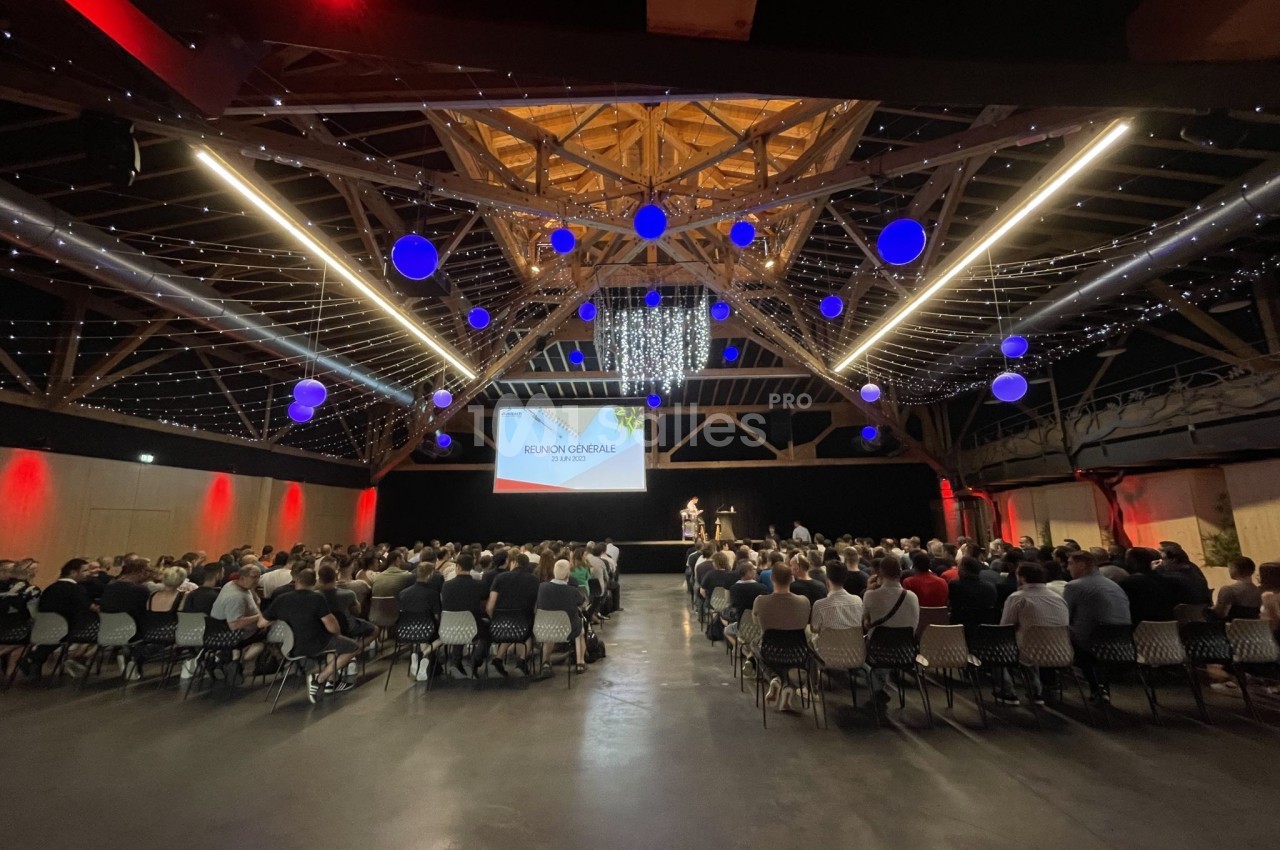 Salle comble avec des participants assis face à une scène, sous une charpente en bois illuminée et des décorations…