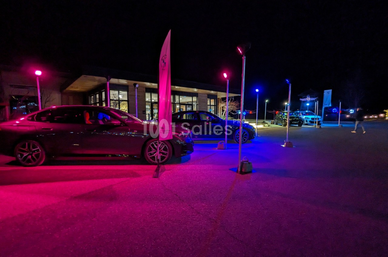Voitures exposées de nuit sur un parking éclairé par des lumières colorées, avec un bâtiment en arrière-plan.