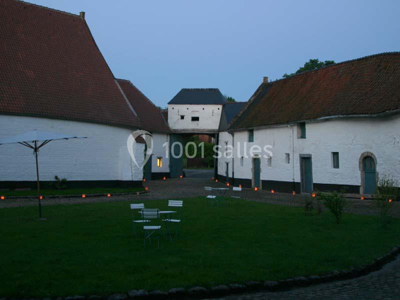 Location salle Court-Saint-Étienne (Brabant wallon) - Ferme de Beaurieux #8 Cour intérieure pavée entourée de bâtiments anciens aux murs blancs et toits en tuiles, éclairée par des bougies au sol.