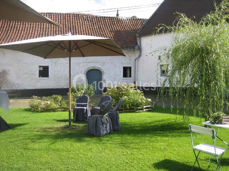 Location salle Court-Saint-Étienne (Brabant wallon) - Ferme de Beaurieux #15 Cour verdoyante avec chaises et parasols, entourée de bâtiments blancs à toit en tuiles rouges.