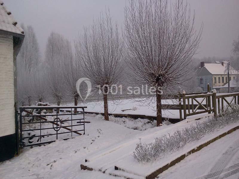 Location salle Court-Saint-Étienne (Brabant wallon) - Ferme de Beaurieux #22 Paysage hivernal avec arbres dénudés, sol enneigé, clôture en bois et maisons en arrière-plan sous un ciel gris.