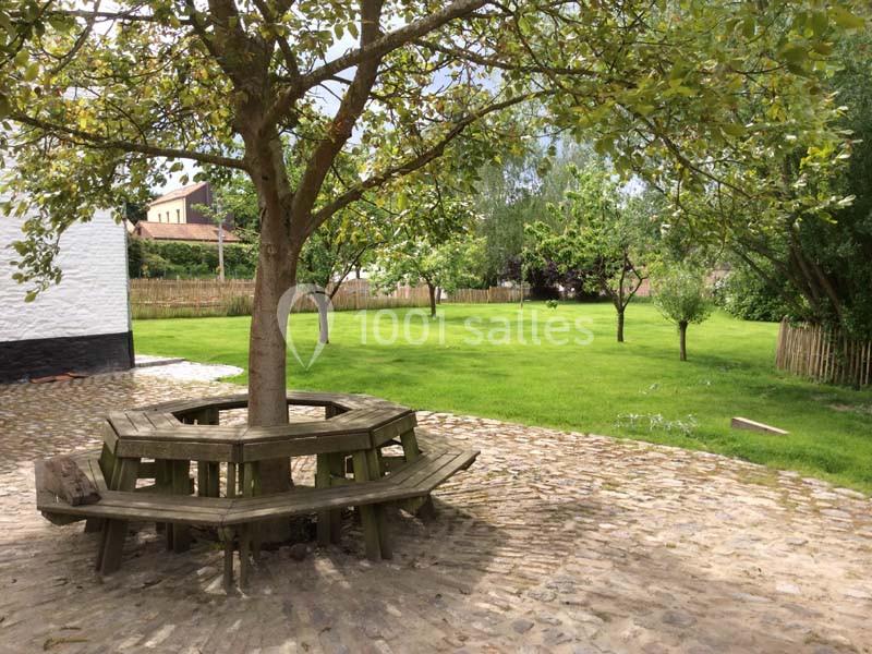 Location salle Court-Saint-Étienne (Brabant wallon) - Ferme de Beaurieux #14 Banc en bois circulaire autour d'un arbre, sur une cour pavée, avec un jardin verdoyant en arrière-plan.