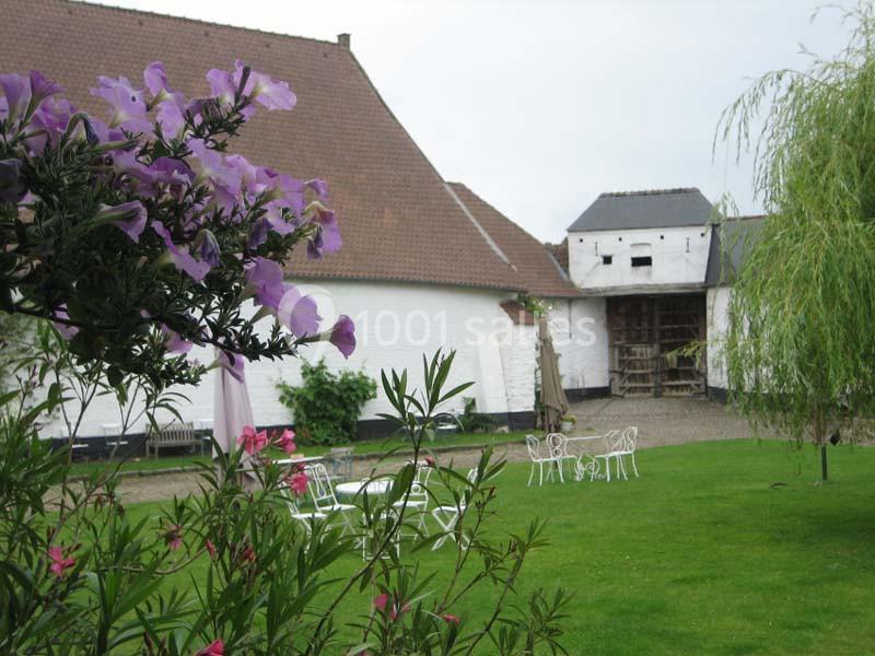 Location salle Court-Saint-Étienne (Brabant wallon) - Ferme de Beaurieux #16 Cour verdoyante avec mobilier de jardin blanc, fleurs au premier plan et bâtiments en briques blanches en arrière-plan.