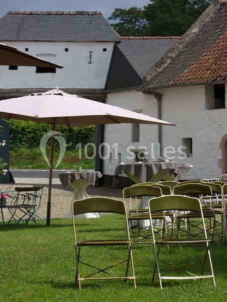 Location salle Court-Saint-Étienne (Brabant wallon) - Ferme de Beaurieux #21 Chaises pliantes disposées sur une pelouse devant des tables et des parasols, près de bâtiments en pierre blanche.