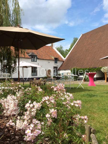 Location salle Court-Saint-Étienne (Brabant wallon) - Ferme de Beaurieux #2 Jardin avec fleurs roses, tables et chaises sous des parasols, devant des bâtiments aux toits en tuiles rouges.