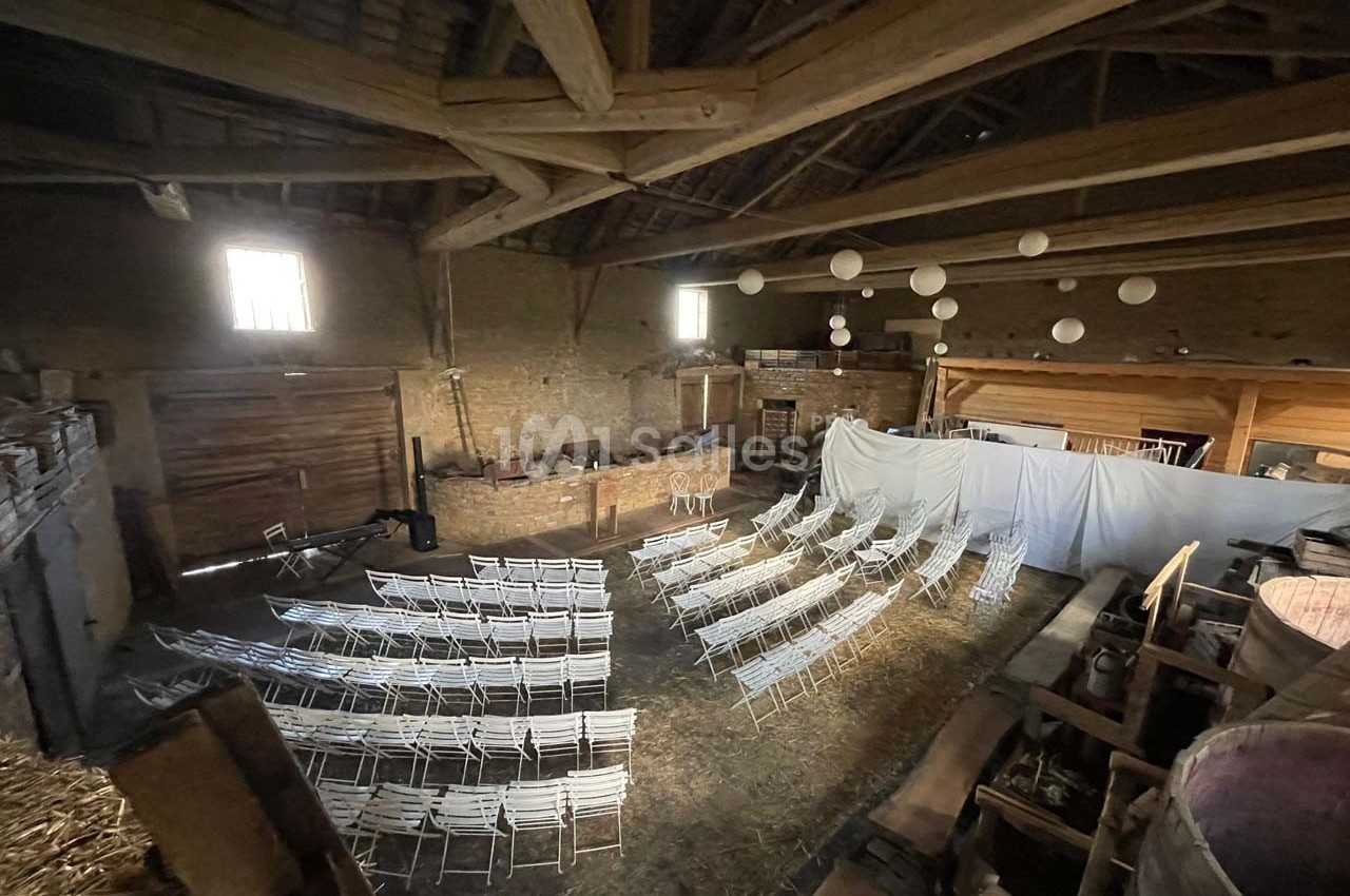 Salle rustique aménagée dans une grange avec des rangées de chaises blanches et des décorations suspendues.